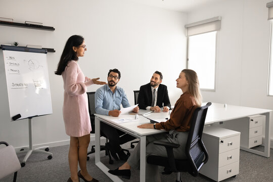 Businesswoman stands near flipchart presenting strategy to colleagues seated at conference table, explaining marketing plan. Business presentation, discussion, collaborative decision making in office