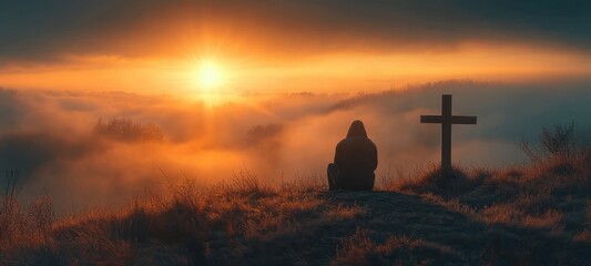 Obraz premium Silhouetted person seated beside a wooden cross on a grassy hill at golden sunrise over misty valleys, evoking quiet contemplation and solemn hope