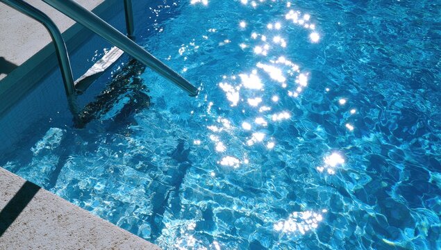 Sunlight dances on rippling blue water of a swimming pool, revealing steps and metal railing
