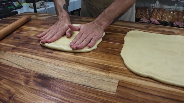 Skilled baker with visible tattoos meticulously rolls out soft, elastic dough on a wooden surface, demonstrating artisan technique in a commercial bakery.