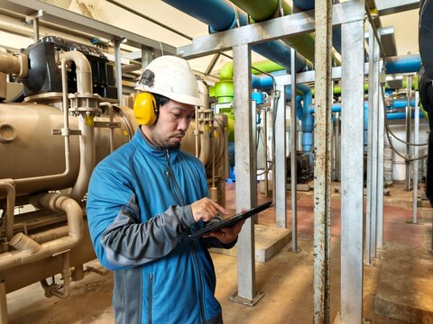 A male industrial engineer in safety equipment focuses on recording machine data on his digital tablet inside a plant