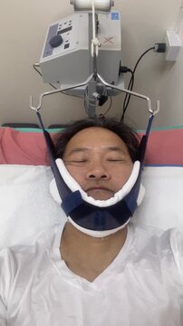 Asian man receiving cervical traction therapy with a medical device in a hospital bed. Treatment for neck pain, pinched nerve, or cervical spine herniated disc.