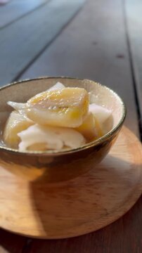 Banana in coconut milk or Kluai Buat Chi. Traditional Thai sweet dessert served in a ceramic bowl on a wooden table.