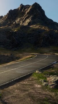 A winding road cuts through a rocky terrain with a large mountain in the background. The scene captures the natural beauty of the landscape under a clear sky.