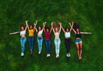 Obraz premium seven young women lying on green grass holding hands raised, smiling and laughing in casual clothes, showing friendship and joy