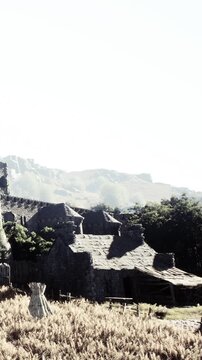 Stone structures sit in a rural setting under clear skies. Nearby mountains rise in the background. There is a rich growth of vegetation around the buildings.