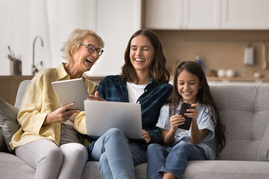 Happy intergenerational women family sitting on sofa with laptop, digital tablet and smartphone spend leisure online, laughing, share memes, enjoy app or funny internet amusements using modern gadgets