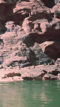 A large rock formation stands next to a flowing river. The water is green and reflects the rocks. People walk along the riverbank enjoying the outdoors.