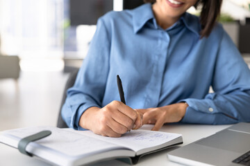 Obraz premium Closeup cropped view of business woman, office employee writing notes in planner, organizes tasks, outlines ideas, updates schedules, and prepares project details, working at modern desk with laptop