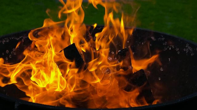 Intense yellow and orange flames consuming wood logs inside a black metal fire pit, with the bright, hot embers glowing against a green grass background during an evening gathering in the backyard