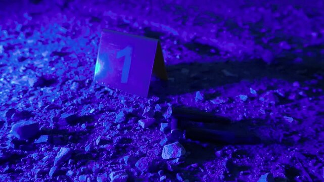 Dramatic crime scene at night showing a yellow evidence marker with the number one next to spent high caliber bullet shells on the ground, illuminated by flashing red and blue police lights