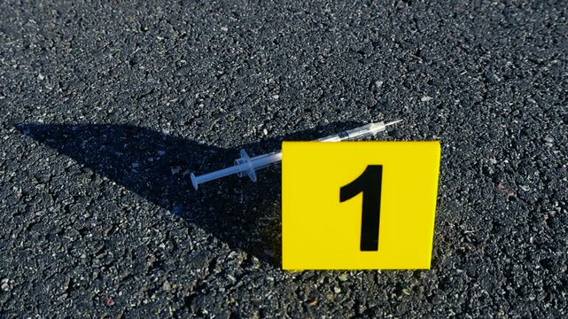 Forensic evidence marker number one placed next to a used hypodermic syringe on an asphalt road during a police criminal investigation, representing drug abuse, overdose, or murder