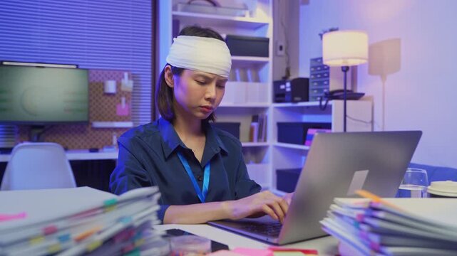 Injured young Asian office worker wearing head bandage while working on laptop in office, health and work pressure concept.
