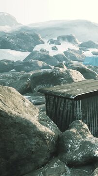 A building is visible among large rocks in a snowy area. The scene shows a mix of gray stones and white snow in a remote environment. The setting appears cold and isolated.