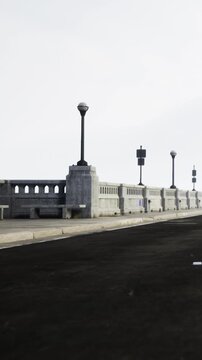People stroll on a walkway at the coast as street lights line the path. The ocean is visible in the background. The sky is gray without clouds.