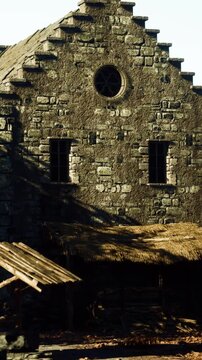 A stone building stands with a thatched roof in daylight. The structure shows signs of age with stone walls and a circular window. Natural light highlights the features of the building.