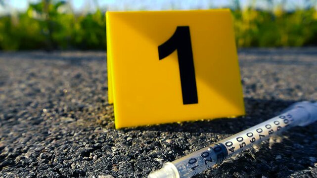 Yellow evidence marker with the number one and a discarded syringe lying on the asphalt pavement, representing a crime scene investigation into drug abuse, addiction, or an overdose death