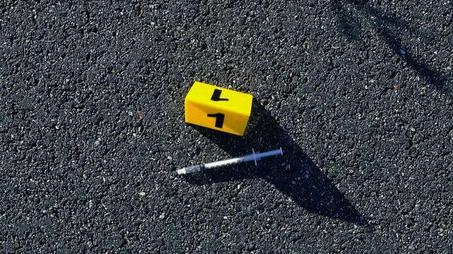 Yellow forensic evidence marker with the number one next to a used syringe on the asphalt, marking a potential piece of evidence at a crime scene related to drug use or an overdose