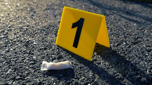 Close up view of a yellow forensic evidence marker number one next to a white nicotine pouch found on the asphalt road, representing the scene of a crime or an ongoing investigation