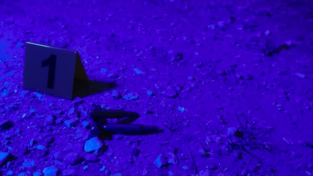 Flashing red and blue police lights illuminating evidence markers next to high caliber bullets on the ground at a crime scene during a night investigation, with the camera panning across the area