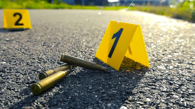 Yellow forensic evidence markers identifying bullet casings found on an asphalt road at a crime scene, with sunlight creating a dramatic shallow depth of field effect in the background