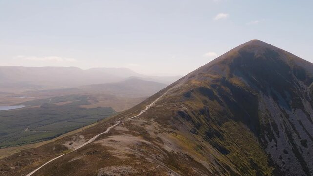 Dramatic drone shot of the Croagh Patrick mountain summit in County Mayo, Ireland. Cinematic view of the holy peak, the small chapel, and pilgrims on the steep trail with Clew Bay in the background.