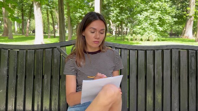 A young brunette woman is studying or working while sitting on a bench in a city park, she is pensive. concept of outdoor studying, learning, or working in a peaceful natural environment.
