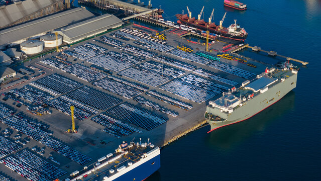 roll-on roll-off (RoRo) vessel docks at a major industrial port facility, surrounded by thousands of new vehicles staged for global export alongside busy container shipping terminals.