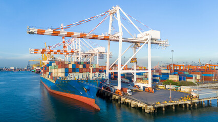 Naklejka premium cargo container ship docked at a commercial port terminal with giant gantry cranes loading freight, representing global maritime logistics, supply chain, and international trade.