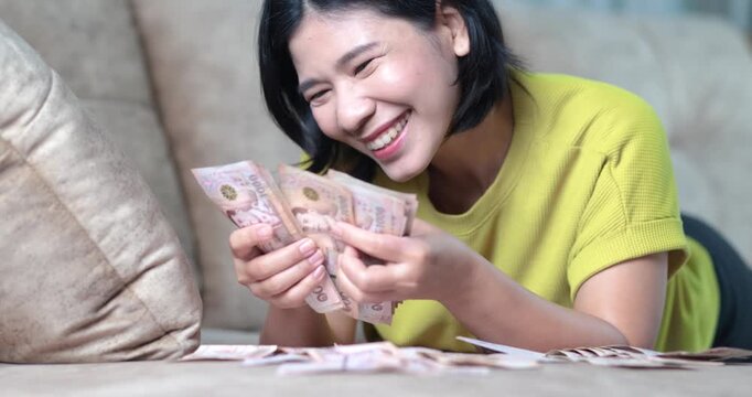 Happy Asian woman counting cash money lying on sofa at home, Thai baht banknote, money, cash, financial success, savings, income, bonus, wealth, and economic stability with cheerful emotion