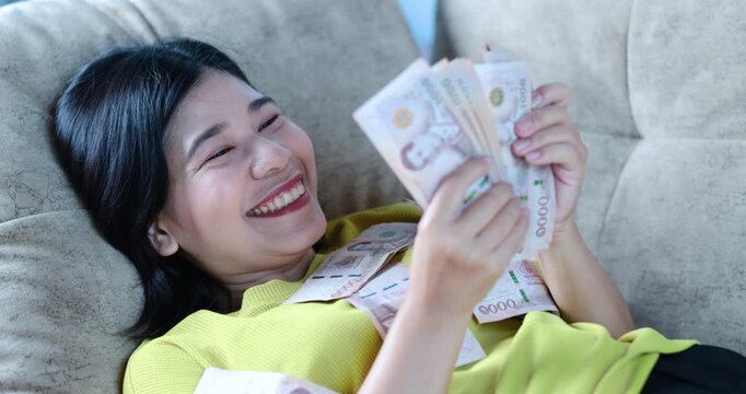 Happy Asian woman counting cash money lying on sofa at home, Thai baht banknote, money, cash, financial success, savings, income, bonus, wealth, and economic stability with cheerful emotion