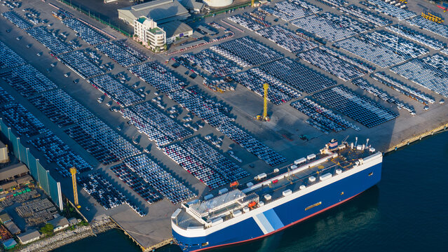 roll-on roll-off (RoRo) vessel docks at a major industrial port facility, surrounded by thousands of new vehicles staged for global export alongside busy container shipping terminals.