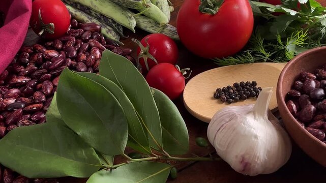 4K slow motion of rustic food composition with raw pinto beans, fresh garlic cloves, ripe red tomatoes and green bay leaves on a wooden table. 