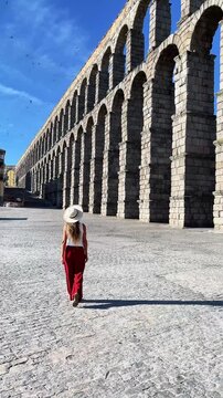Back view of female tourist in red trousers and straw hat walking by ancient Roman aqueduct in Segovia Spain