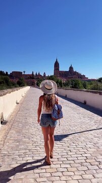 Back view of female tourist with backpack walking across old stone bridge towards Salamanca cathedral in Spain