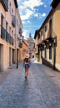 Back view of young female tourist in shorts and straw hat walking in street in Segovia, Spain