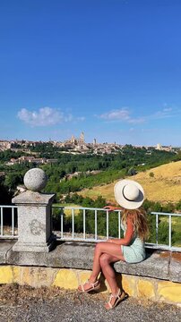Young woman in dress and straw hat sitting on stone wall looking at Segovia city skyline in Spain