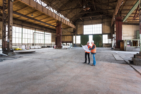 Two men with plan wearing safety vests talking in old industrial hall