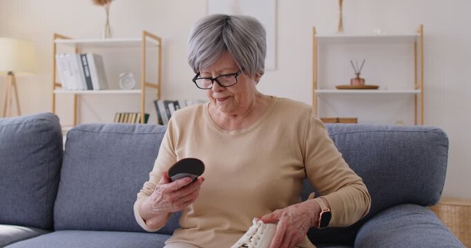 Senior elderly woman checks orthopedic insole for foot pain. She sits on a sofa, checking a shoe on flatfoot to boost comfort, mobility, and health. Concept foot relief and feet comfort at home.