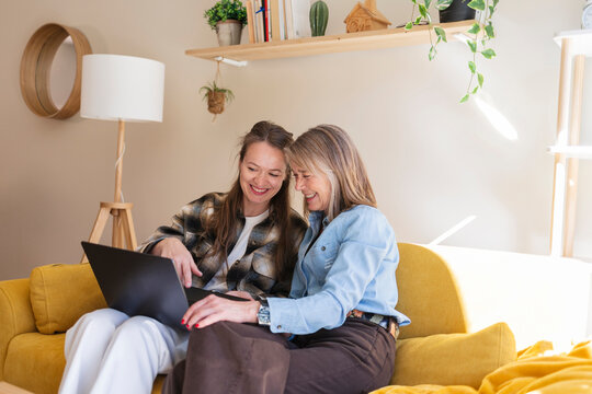 Happy mother and daughter laugh and share a laptop on sofa at home