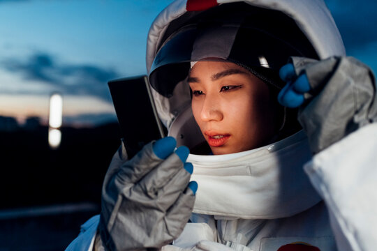 Female astronaut looking at smart phone while holding helmet at night