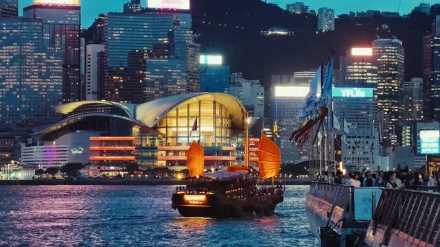 A bustling nighttime scene in Hong Kong featuring a traditional junk boat gliding across the waterfront