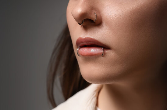 Pretty young woman with nose and lip piercing on grey background, closeup