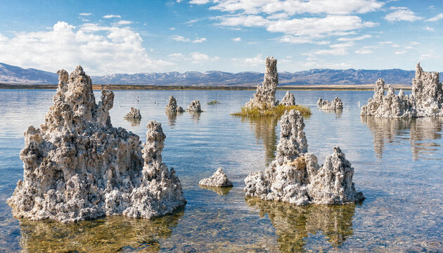 Tufa towers rising from the calm water of mono lake with a backdrop, of distant snow-capped, mountains under a, bright blue sky, with clouds