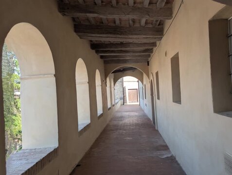 Corridoio porticato con travi in legno e pavimento in cotto, illuminato da archi laterali che creano giochi di luce e ombra. Atmosfera storica