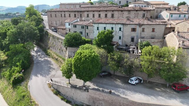 Veduta di San Vito sul Cesano con strada panoramica accanto a muro in pietra, edifici storici in pietra chiara e colline marchigiane sullo sfondo