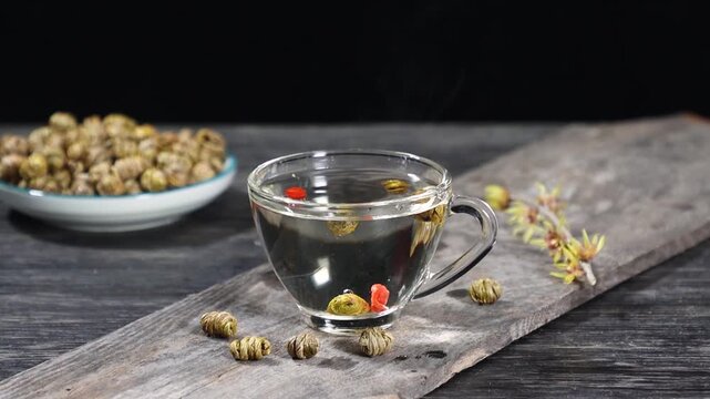Slow motion of dendrobium orchid herb dropping into a glass teacup with goji berries on a rustic wooden table.