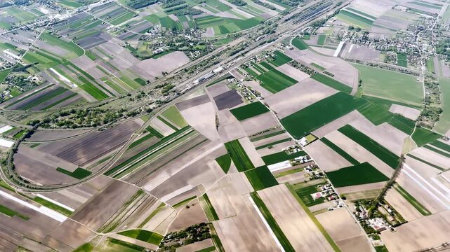 Aerial view of patchwork farmland with green and pale crop fields, railway corridor, country roads and small villages in bright daylight.