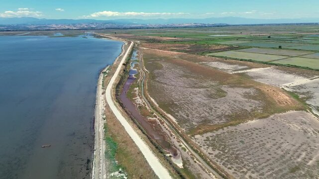 Cinematic drone view flying along a scenic coastline road separating a calm blue sea from vast delta wetlands and green rice fields.