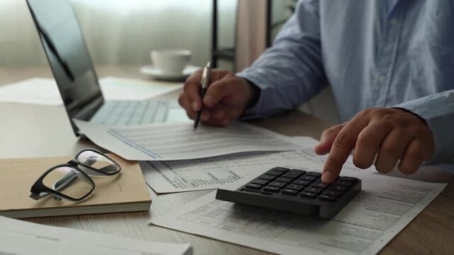 man Hands review financial reports with a calculator and pen beside a laptop, checking figures on printed documents for accounting analysis, budgeting records, and business finance workflow.